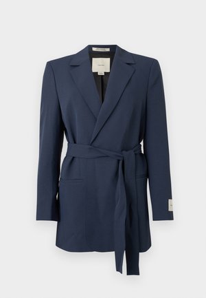 Marineblå blazer med belte, hakket slag, lange ermer og lommer med åpning foran, vist mot en hvit bakgrunn.