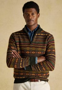 Kötött pulóver színes geometriai mintákkal, földszínekkel, mint zöld, narancs és piros, negyedcipzáros gallérral és bordás mandzsettákkal.