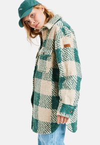 Een groen en crèmekleurig plaid shacket gemaakt van zachte, gestructureerde stof, met een kraag, twee voorzakken en een logopatch op de mouw.