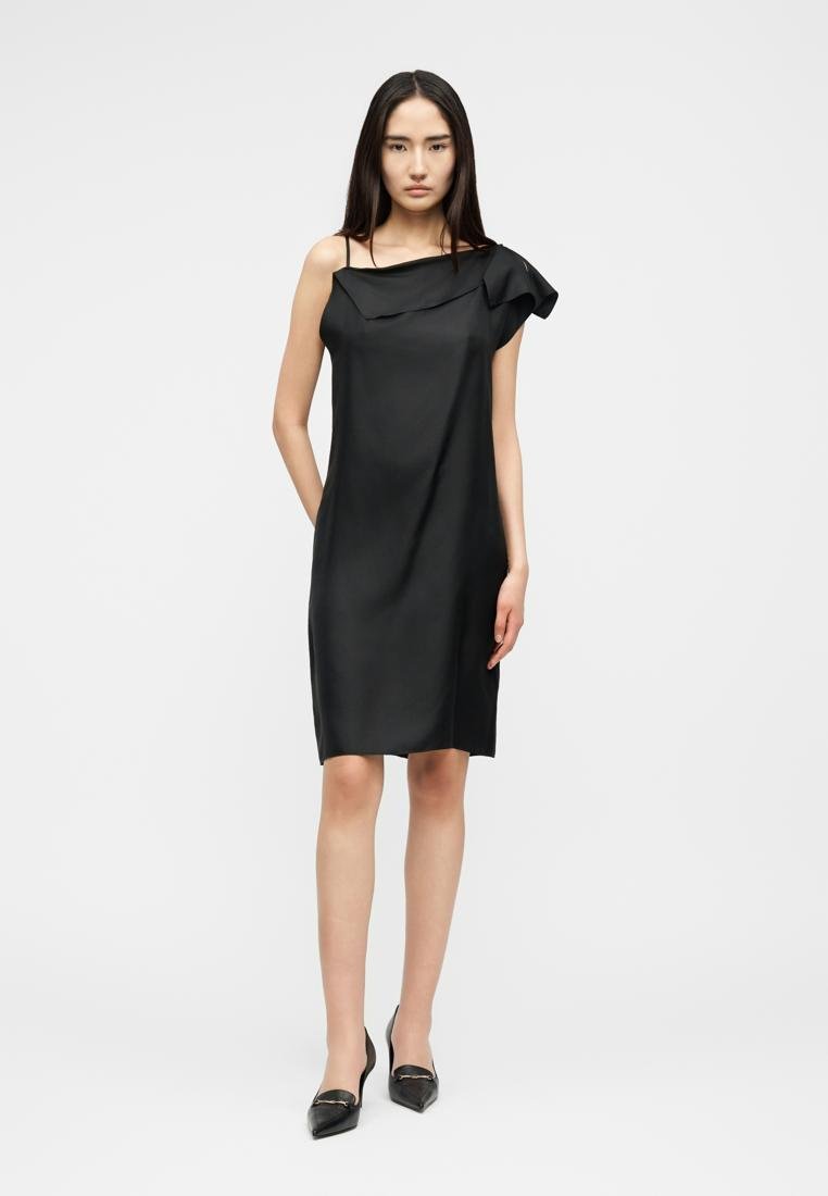 Femme debout portant une robe noire asymétrique mi-longue avec une épaule à volants et des escarpins noirs à bout pointu sur fond blanc.