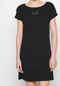 Czarna bawełniana sukienka z podwiniętymi rękawami i dekoltem okrągłym. Posiada złote logo "EA7" z kryształkami z przodu. Prosty, luźny fason.