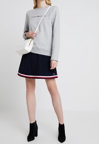 Femme portant un sweat-shirt gris Tommy Hilfiger, une jupe marine avec des bordures rouge, blanc et bleu, des bottines noires et un sac en bandoulière blanc.