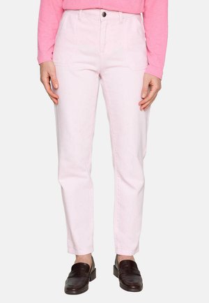 Femme portant un pantalon droit rose clair, un haut à manches longues rose et des mocassins marron foncé, debout avec les mains détendues le long du corps.