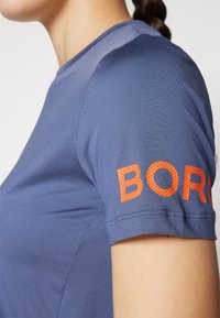 Rövid ujjú, kék sportpóló sima textúrával, bal karján narancssárga perforált "BORG" logóval.