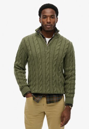 Olive green cable knit sweater with a zip collar, featuring ribbed cuffs. Layered over a plaid flannel shirt with blue and brown accents.