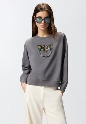 Donna con felpa grigia scuro con design di uccello colorato in paillettes e pantaloni crema, mani nelle tasche e grandi occhiali da sole neri.