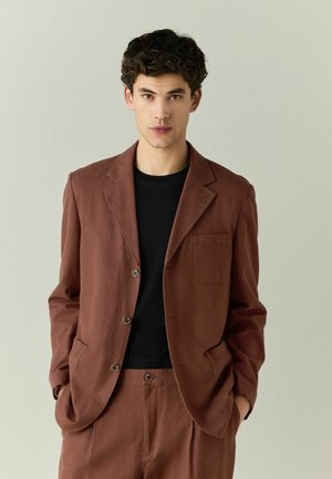 Young man wearing a brown suit with a black shirt underneath, standing with hands in pockets against a plain light background.