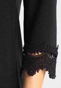 Svart långärmad tröja med intrikat svart spetsdetalj vid ärmslutet på en persons arm mot en vit bakgrund.