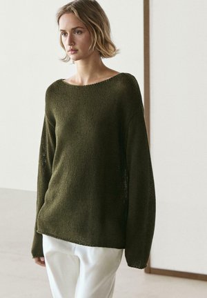 Žena v olivově zeleném pleteném svetru s dlouhými rukávy a bílých kalhotách stojí na jednobarevném světle pozadí.