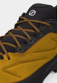 Μαύρο και κίτρινο παπούτσι πεζοπορίας με κορδόνια κίτρινου και μαύρου με σχέδιο, σουέτ υφή και λευκό λογότυπο στη μαύρη γλώσσα.