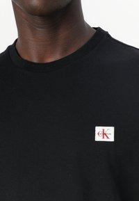 Donkerhuidig persoon draagt een zwart T-shirt met ronde hals en een klein wit Calvin Klein-logo op de linkerborst.