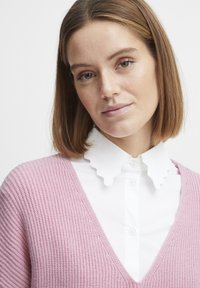 Roza pletena jopica z globokim V-izrezom, složena preko bele majice s valovitim ovratnikom. Gladko teksturo, dobro definirani vzorci.