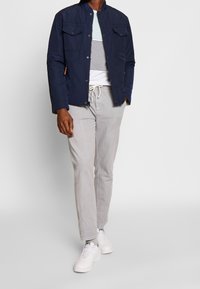 Giacca leggera blu navy con due tasche sul petto, abbinata a una camicia a righe a maniche corte, pantaloni grigi chiari e sneaker bianche.