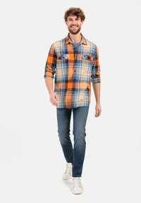 Chemise en flanelle à carreaux orange, bleu et beige avec deux poches poitrine ; associée à un jean bleu et des baskets blanches. Le modèle est en train de marcher.