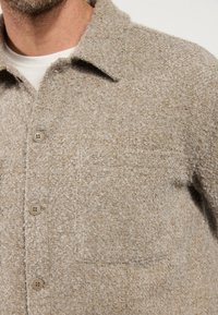 Brązowa koszula zapinana na guziki z teksturowanej tkaniny z kołnierzykiem, posiadająca kieszeń na piersi i beżowe guziki.