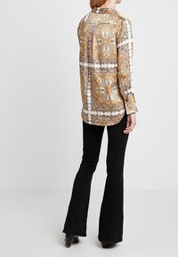 Långärmad skjorta i guld- och vit paisley-mönster, tillverkad av ett slätt tyg, med krage med knappar och rak snitt, kombinerad med utsvängda svarta byxor.