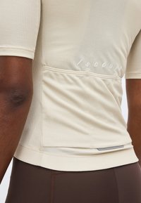 Μπεζ κυκλική φανέλα με κοντά μανίκια και υφή, με πίσω τσέπες και ανακλαστική ταινία, φορεμένη με σκούρα καφέ εφαρμοστά κάτω μέρη.