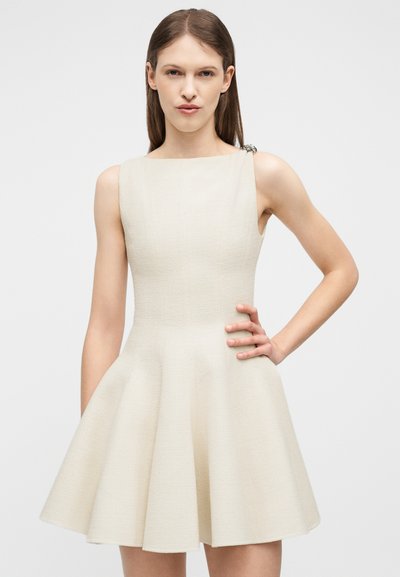 Γυναίκα με ίσια καστανά μαλλιά, φοράει αμάνικο ελεφαντόδοντο φόρεμα με υφή, εφαρμοστό στη μέση και φαρδαίνει προς τα κάτω, με ασημένια πόρπη στον αριστερό ώμο, χέρι στη μέση.