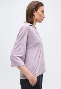 Kvinne iført en løs, lys lilla, knappet topp med trekvartlange ermer, stående i profil med en hånd i lommen på mørke bukser.