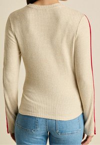Beżowa bluzka z długim rękawem w prążki z czerwonymi paskami po bokach. Materiał jest teksturowany, ma dopasowany krój i zaokrąglony dekolt.