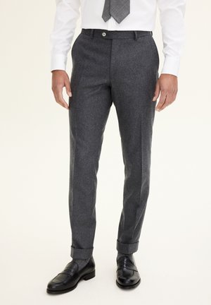 Pantaloni di lana grigi con vestibilità sartoriale, davanti piatto e orli a doppio risvolto. Indossati con una camicia bianca e una cravatta a fantasia, abbinati a mocassini neri.