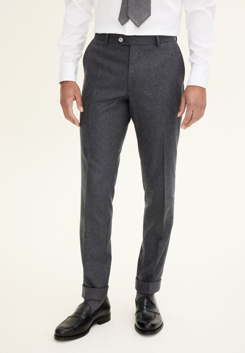 Szare wełniane spodnie o dopasowanym kroju, z płaskim przodem i mankietami. Noszone z białą koszulą i wzorzystym krawatem, w zestawieniu z czarnymi mokasynami.