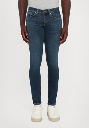 Man in slim-fit donkerblauwe jeans met vijf zakken, witte sneakers en een lichtgrijs shirt, staand tegen een witte achtergrond.