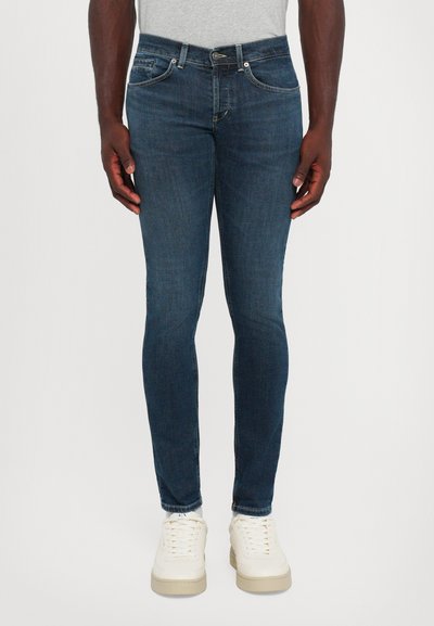 Man in slim-fit donkerblauwe jeans met vijf zakken, witte sneakers en een lichtgrijs shirt, staand tegen een witte achtergrond.