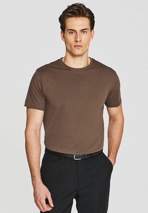 Młody mężczyzna z krótkimi brązowymi włosami, ubrany w gładki brązowy T-shirt z krótkim rękawem i czarne spodnie z czarnym plecionym paskiem, stojący na jednolitym tle.