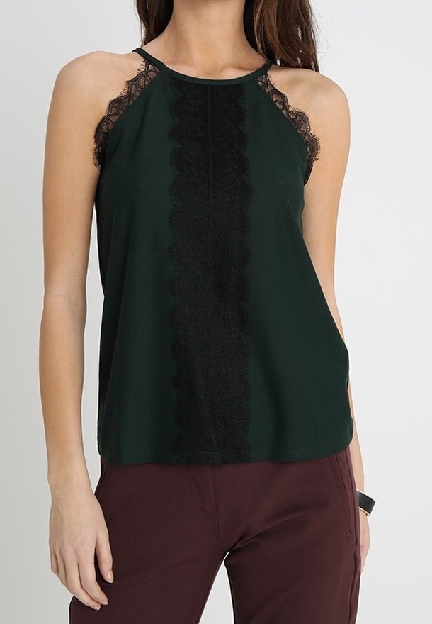 Chemisier sans manches vert foncé avec bordure en dentelle noire sur le devant et les épaules, porté avec un pantalon bordeaux foncé.