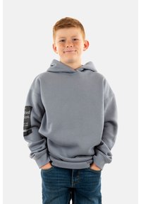 Sudadera gris con un bolsillo delantero, corte oversized y puños acanalados. Presenta texto y un diseño gráfico en la manga izquierda. Se lleva con jeans azules.