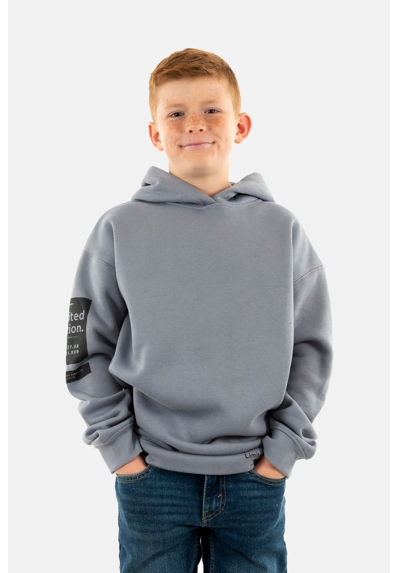 Sudadera gris con un bolsillo delantero, corte oversized y puños acanalados. Presenta texto y un diseño gráfico en la manga izquierda. Se lleva con jeans azules.