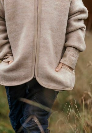 Beige fleecetakki vetoketjulla, jossa suuret taskut ja resorit hihansuissa, puettuna tummansiniin cargo-housuihin nurmikkoisessa ulkotilassa.