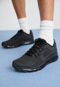 Sapatos atleticos pretos com parte superior em malha, colarinho acolchoado, atacadores pretos sólidos e uma sola espessa e almofadada. Usados com meias brancas caneladas.