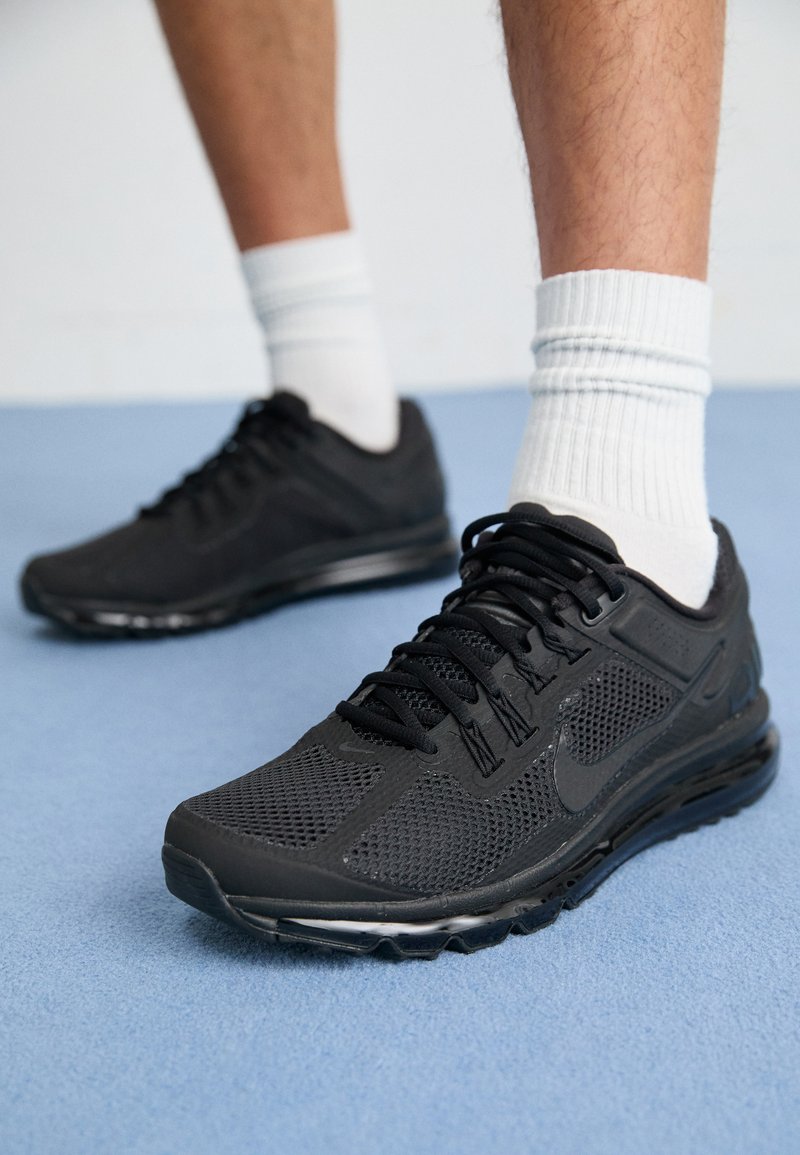 Zapatillas deportivas negras con parte superior de malla, cuello acolchado, cordones negros sólidos y suela gruesa y acolchada. Llevadas con calcetines blancos acanalados.