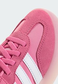 Růžová sportovní obuv vyrobená z nubuku a syntetického materiálu, s texturovaným povrchem, výraznými bílými pruhy a kulatým designem špičky.