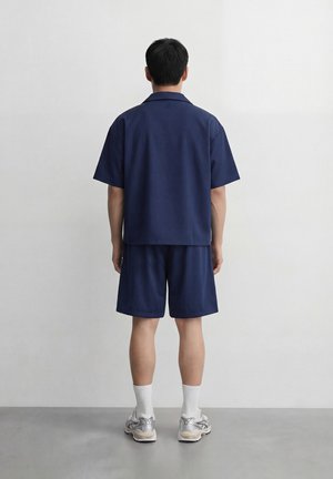 Man standing facing a white wall wearing a navy blue short-sleeve shirt, matching shorts, white socks, and silver athletic shoes.