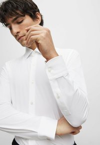 Camisa blanca de manga larga con un collar clásico, cierre de botones en la parte frontal y detalles en los puños. El material parece suave con un ligero brillo.