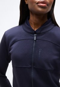 Κοντινό πλάνο ενός ατόμου που φοράει μια μπλε ρεβένζεια ζακέτα με φερμουάρ και το λογότυπο Calvin Klein στο στήθος, με ριμπ κοντό γιακά.