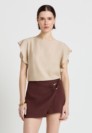 Woman wearing a beige short-sleeve blouse and a dark brown wrap skirt with decorative metal rings against a plain background.