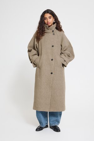 Une femme porte un long manteau taupe à col montant et à boutons foncés, associé à un jean bleu et à des chaussures noires, sur un fond de studio uni.