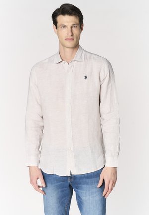 Uomo con capelli scuri che indossa una camicia beige chiaro a maniche lunghe con bottoni e jeans blu, in piedi davanti a uno sfondo bianco semplice.