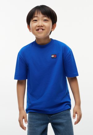Jeune garçon souriant portant un t-shirt bleu Tommy Hilfiger et un jean bleu, debout devant un fond blanc uni.