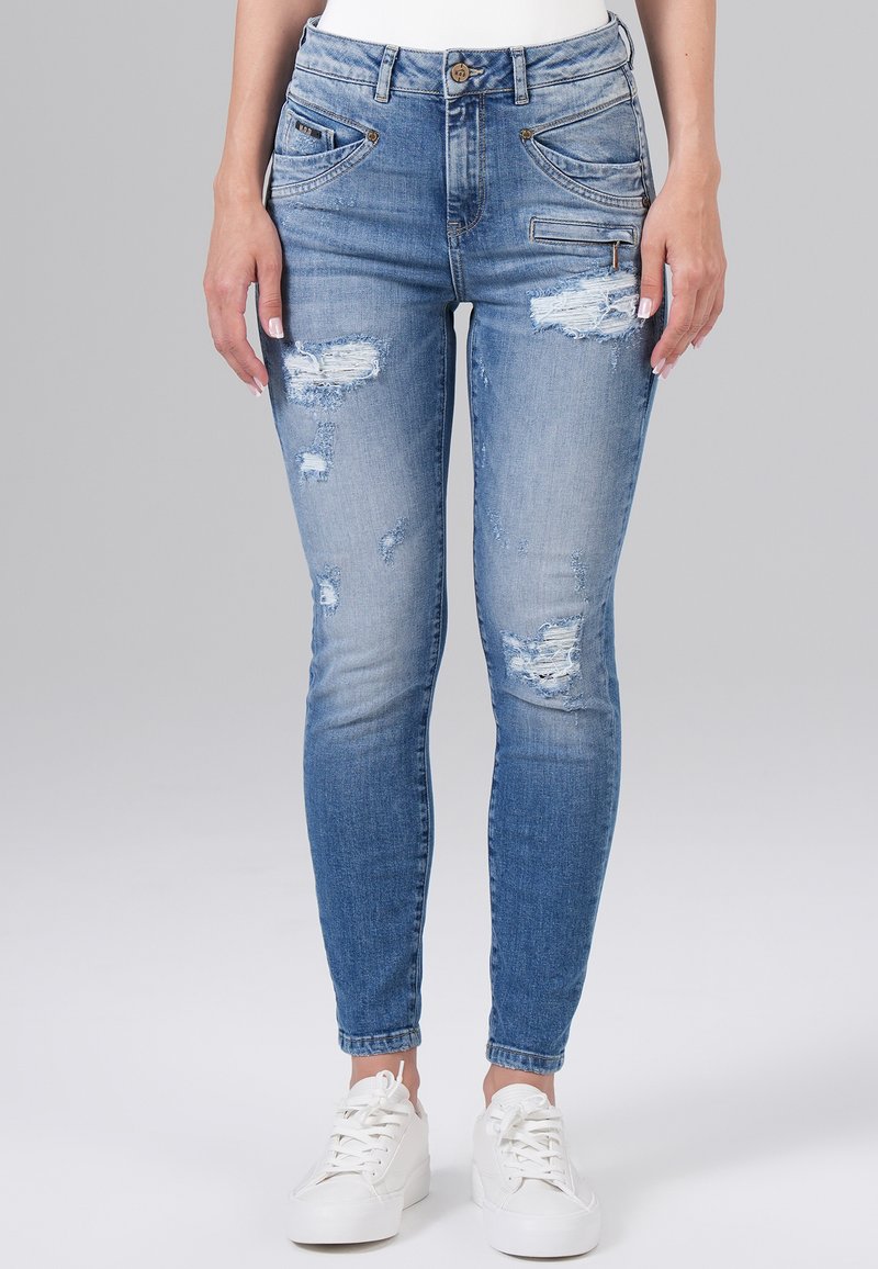 Jeans skinny en denim bleu clair avec des patchs déchirés et des zones décolorées. Comprend cinq poches et une fermeture à bouton. Associés à des baskets blanches.