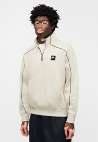 Ανοιχτό μπεζ φούτερ με φερμουάρ, μαύρο περίγραμμα, μπροστινή τσέπη και μικρό μαύρο λογότυπο Nike Air. Κατασκευασμένο από υφασμάτινη ύλη με υφή, με μακριά μανίκια.