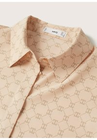 Blouse beige à col avec un motif géométrique subtil de formes entrelacées, étiquette de tissu indiquant la marque et la taille à l'intérieur de l'encolure.