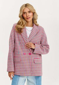 Žena s blond vlasy, oblečená v růžovo-modro-žlutém kostičkovaném saku s jasně růžovými knoflíky, pod kterým má bílé tričko a světle modré džíny.