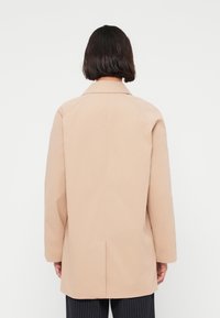 Personne aux cheveux courts et foncés portant un long manteau beige, vue de dos sur un fond clair uni.