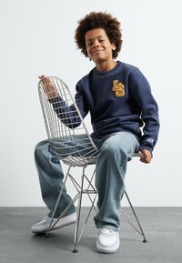 Námornícky sveter s žltou výšivkou "LS", džíny, sedí na striebornej drôtenej stoličke. Biele tenisky s modrými akcentmi. Hladké pozadie.