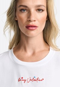 T-shirt en coton blanc avec broderies rouge et bleue portant le texte "sois mon Valentine". Col rond, texture douce, coupe décontractée.